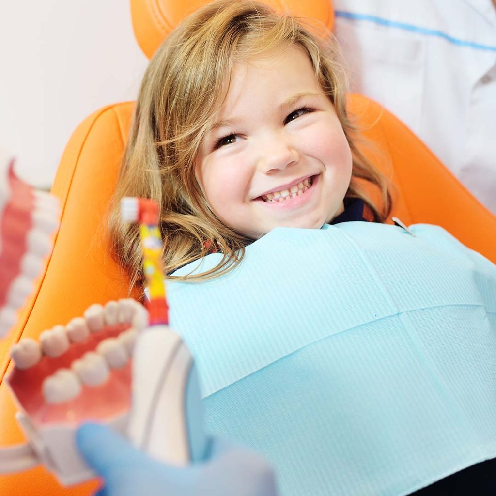 pediatric dentist shows how to properly brush her teeth to a little girl who is sitting in an orange dental chair and smiling.